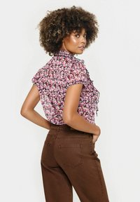 Rožinė gėlių bluzonė su raukiniais ir trumpomis rankovėmis, derinama su rudomis aukštu liemeniu džinsais, matoma iš šono. Laižyta medžiaga.