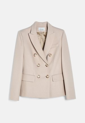 Kvinde med glat brunt hår iført en beige dobbeltradet blazer og bukser med vide ben, accessorized med guldsmykker, stående med hænderne bag ryggen.