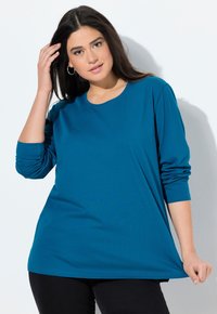 Langarm-T-Shirt in Teal aus einem weichen, leichten Stoff mit Rundhalsausschnitt und lockerer Passform; getragen mit schwarzen Hosen.