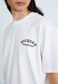 Hvit bomull T-skjorte med et svart og rosa grafisk logo med teksten "DICKIES WORKWEAR," kort erm, og rund hals.