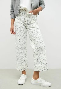 Witte bloemenbroek met marineblauwe patronen, wijdvallend ontwerp en textuur van de stof. Gecombineerd met witte sneakers en een lichtgrijze cardigan.