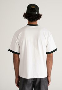 Bărbat cu păr creț, purtând un tricou alb cu finisaje negre și o pălărie închisă la culoare cu inscriptionarea "MODERN YUPPIES," întors cu spatele, pe un fundal simplu.