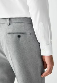 Pantalones grises a medida con una textura suave, que presentan un bolsillo trasero con botón. Se vislumbra parcialmente una camisa blanca de manga larga.
