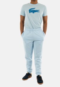 Maglietta in cotone azzurro chiaro con logo di un coccodrillo blu, abbinata a pantaloni da tuta azzurro chiaro con vita elastica e tasche. Scarpe da ginnastica nere.
