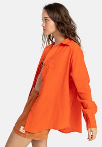Camicia arancione con bottoni, dalla vestibilità morbida e maniche lunghe, abbinata a pantaloni corti marroni. La camicia ha una superficie liscia e un colletto classico.