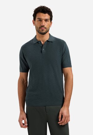 Man met krullend haar, gekleed in een donkergroen poloshirt met korte mouwen en een bijpassende donkere broek, staand tegen een effen achtergrond.