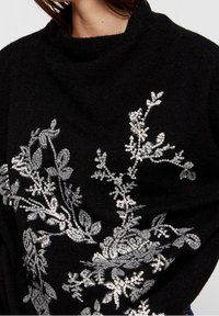 Zwarte trui met een hoge halslijn, versierd met een textuur van florale ontwerpen in zilveren pailletten, wat een contrast vormt met de stof.