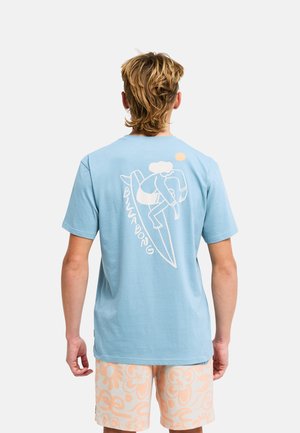 Osoba nosząca jasnoniebieski t-shirt z białą grafiką deski surfingowej i tekstem "Billabong" na plecach, w połączeniu z jasnymi, kwiatowymi szortami.