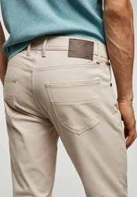 Beige broek met een gladde textuur, voorzien van twee achterzakken met gestikte randen en een bruine leren patch op de tailleband.