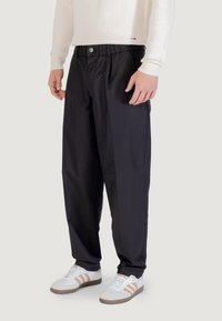 Czarne, plisowane spodnie z elastycznym pasem i zapięciem na guzik. W zestawieniu z białymi trampkami z brązowymi akcentami, na neutralnym tle.