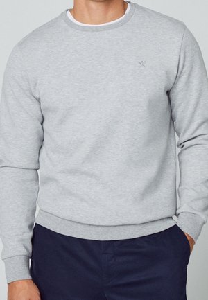 Mann trägt hellgraues Sweatshirt mit Rundhalsausschnitt und kleinem besticktem Logo auf der Brust, kombiniert mit dunkelblauen Hosen, Hände in den Taschen.