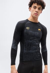 Νεαρός άνδρας που φοράει μαύρο αθλητικό πουκάμισο με μακριά μανίκια, με σκούρα γκρι σχέδια και χρυσά λογότυπα ζώων, στέκεται μπροστά σε ένα απλό φωτεινό φόντο.