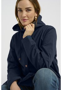 Veste à capuche bleu marine en tissu texturé, avec une fermeture frontale à boutons-pression de teinte cuivrée et une capuche ajustable avec cordon de serrage.