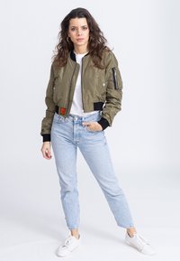 Giubbotto bomber verde oliva con tasca con zip, polsini e orlo a coste. Abbinato a jeans blu chiaro e sneakers bianche.