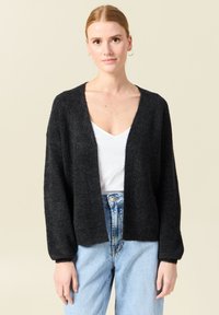 Svart, stickad cardigan med V-ringning, långa ärmar och en avslappnad passform, bärs över en vit topp och blå högmidjade jeans.