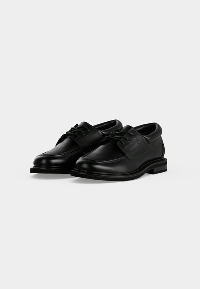 Paire de chaussures de ville en cuir noir à lacets, bout arrondi et talon bas sur fond clair uni.