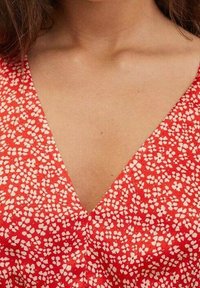 Rotes V-Ausschnitt-Kleid mit kleinen weißen Blumenmustern. Der Stoff hat eine glatte Textur und einen lockeren Schnitt um den Ausschnitt und die Schultern.