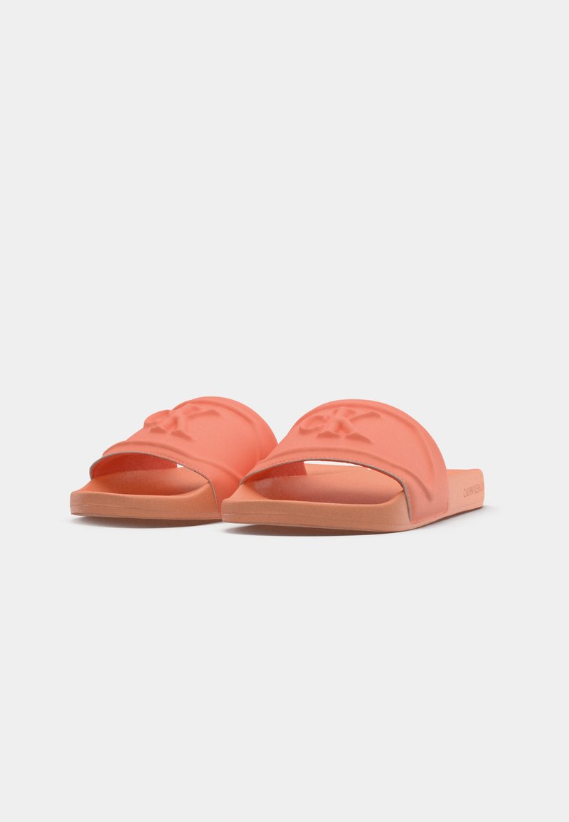 Chanclas de coral con una textura suave y mate, correas anchas con un logo en relieve y una suela plana y contorneada.
