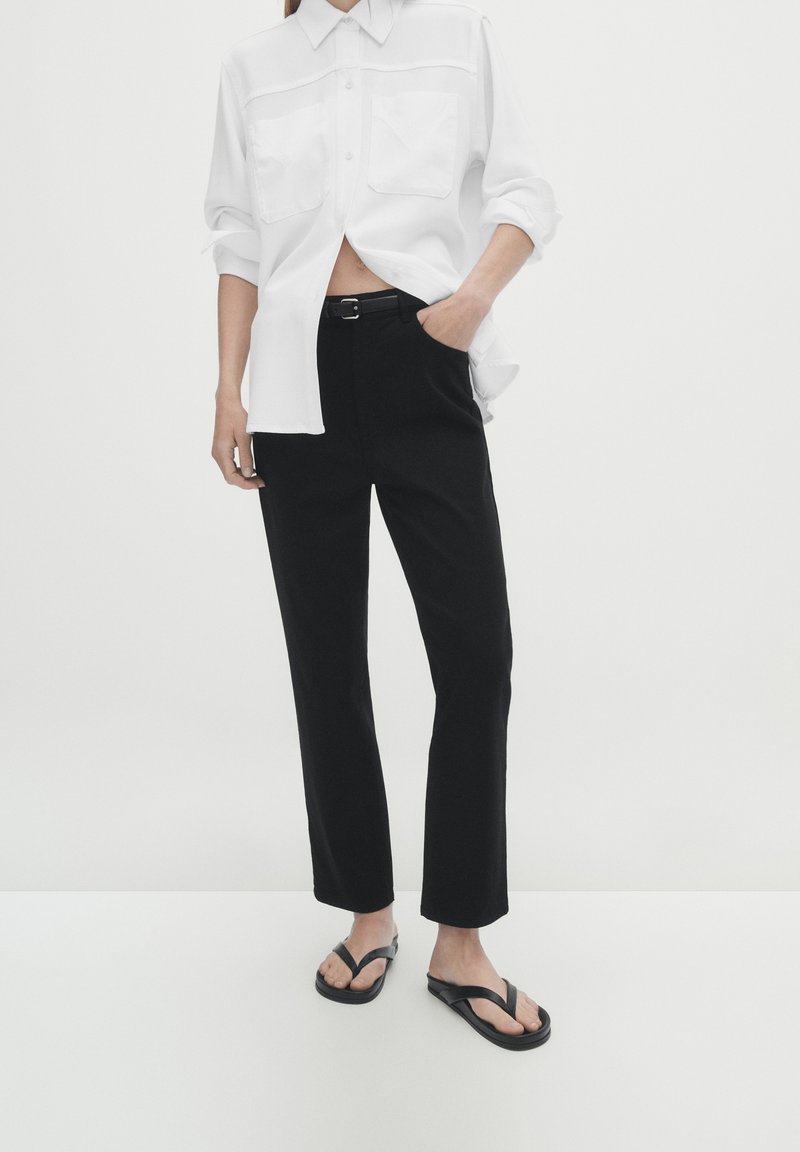 Personne portant une chemise blanche à boutons avec les manches retroussées, un pantalon noir ceinturé et des sandales flip-flop noires, debout devant un fond uni.