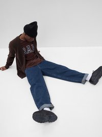 Brun strikket cardigan, mørkebrune t-skjorte med "GAP" logo, blå løse jeans, svart lue, svarte sko, sittende mot en grå bakgrunn.