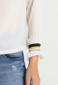 Weiße Bluse mit einem Rüschenbund, die marineblaue und gelbe Streifen aufweist. Glatter Stoff, semi-transparenter Stoff und ein verkürztes Design.