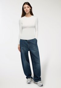 Giovane donna con lunghi capelli castani che indossa un top fitted bianco a maniche lunghe, jeans scuri larghi e scarpe da ginnastica argentate, in piedi con una mano in tasca.