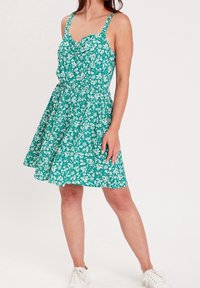 Robe fleurie verte avec bretelles ajustables, corsage cintré et jupe évasée. Le tissu est léger, orné de motifs floraux blancs.