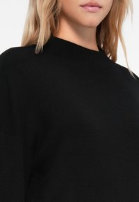Fekete kötött pulóver magas nyakkal és laza fazonnal. Simára kidolgozott textúra, hosszú ujjak és minimalista design jellemzi.