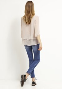 Blusa decolor beige con mangas tres cuartos, diseño en capas. Llevada con jeans ajustados azules y zapatos negros de cordones con suela gruesa.