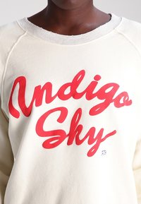 Sweat-shirt crème avec un texte cursif rouge vif "Indigo Sky" sur le devant. Texture douce avec un col rond côtelé et des manches raglan.