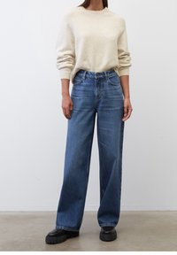 Jasno beżowy sweter dżersejowy z prążkowanymi mankietami, w połączeniu z wysokim stanem szerokimi niebieskimi dżinsami. Masywne czarne buty na platformie dopełniają całość.