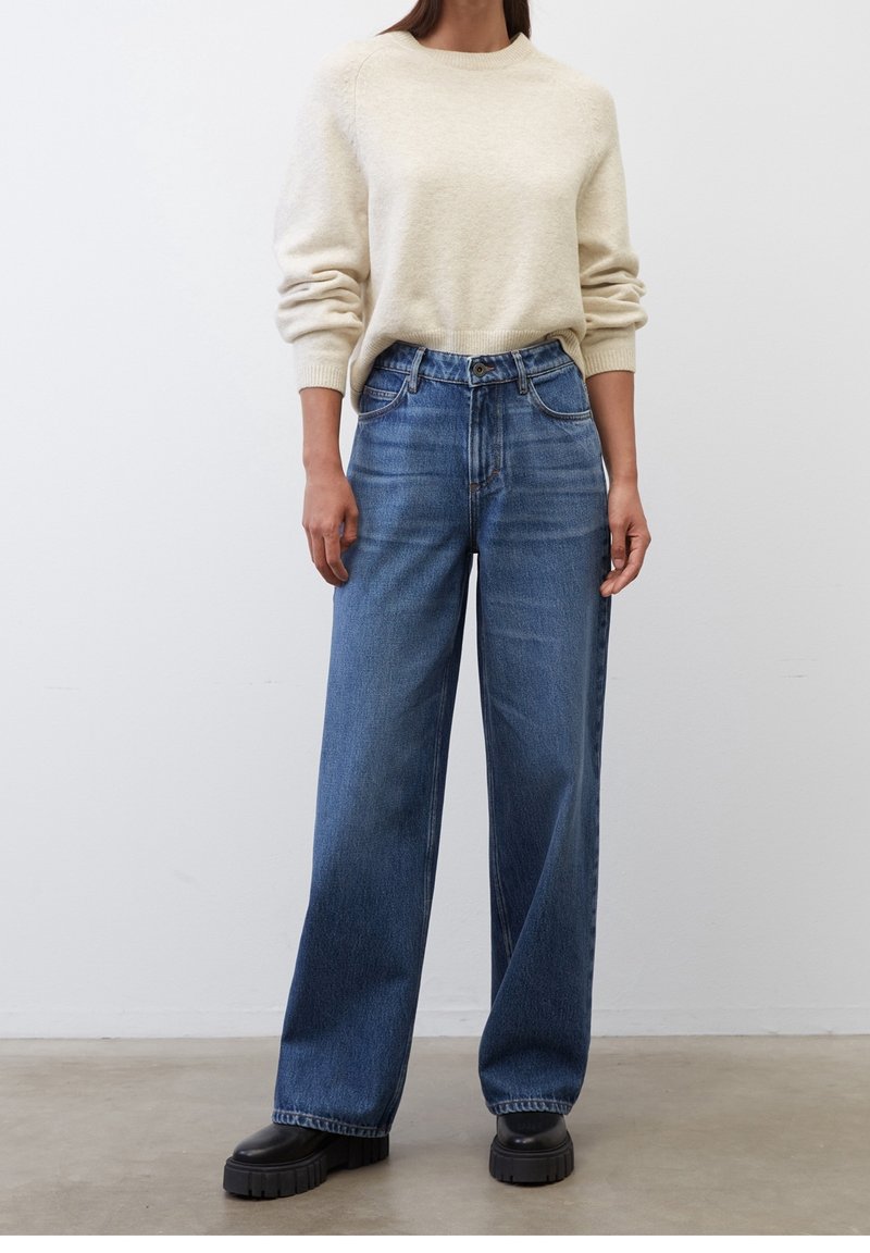 Jasno beżowy sweter dżersejowy z prążkowanymi mankietami, w połączeniu z wysokim stanem szerokimi niebieskimi dżinsami. Masywne czarne buty na platformie dopełniają całość.