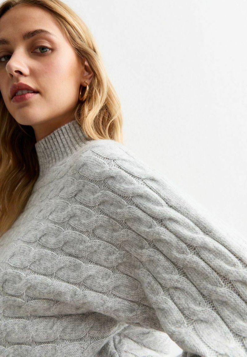 Maglione a trecce di colore grigio chiaro, con colletto alto e texture a costine. Motivo a maglia spessa con tessuto morbido e caldo.