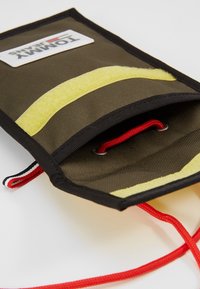 Busta in tessuto khaki con striscia in velcro gialla e cordino rosso; presenta bordi neri e occhiello metallico per una chiusura sicura. Patch del logo visibile.