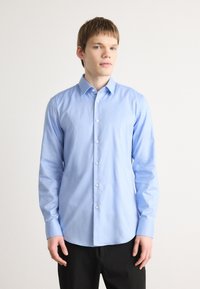 Camisa de botones azul claro en un tejido suave, con mangas largas, cuello clásico y botones blancos, combinada con pantalones oscuros.