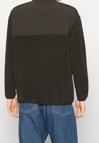 Mörk olivgrön långärmad skjorta, tillverkad av mjukt tyg. Har en kontrasterande slät panel på den övre ryggen och rundad nederkant.