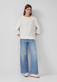 Witte, gestructureerde trui met een ronde halslijn en 3/4 mouwen, gecombineerd met lichtblauwe wijde jeans en witte sneakers.