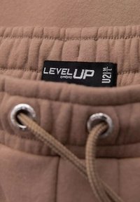 Pantaloni jogger marroni con vita elastica e cordino. Presentano un'etichetta con logo "LEVEL UP" e occhielli in argento per il cordino. Texture morbida.