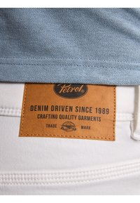 Tejido azul claro en la parte superior con un acabado texturizado. Abajo, una etiqueta de cuero marrón presenta texto negro, indicando la marca y detalles del producto. Se muestra tela blanca por debajo.