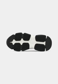 Semelle de chaussure de sport noire et blanche avec des sections de grip en caoutchouc segmentées et un motif en chevrons, vue de dessous.