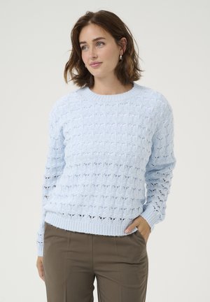 Helles blaues Strickpullover mit einem strukturierten Maschenmuster, Rundhals-Ausschnitt, gerippten Bündchen und Saum. Über braunen Hosen getragen.