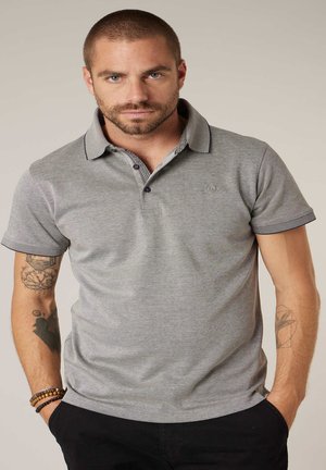 Homme aux cheveux courts et à la barbe portant un polo gris et un pantalon noir, montrant des tatouages sur les deux bras, les mains dans les poches.