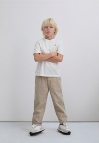 Bílé bavlněné tričko s krátkými rukávy, jemný červený detajl loga, doplněné béžovými volnými kalhotami a bílými teniskami s tmavými akcenty.