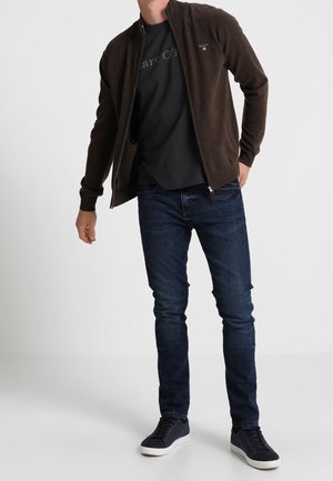 Brązowa kurtka na zamek z ciemnym t-shirtem, niebieskimi dżinsami i ciemnymi trampkami. Gładki materiał z luźnym krojem. Minimalistyczny design bez wyraźnych wzorów.