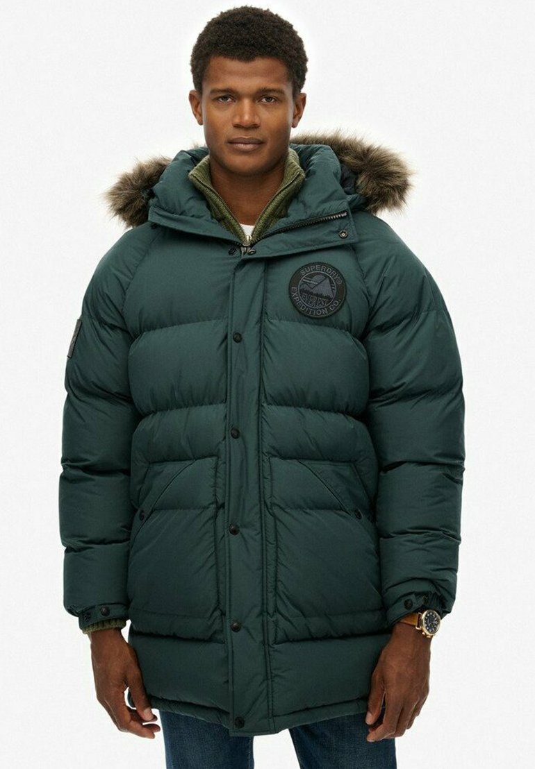 Groene pufferjack met capuchon, voorzien van een bontafwerking, ritsluiting en een patch op de borst. Inclusief twee voorzakken en een gestikte textuur.