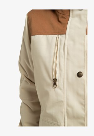 Beige jas met bruine schouderaccenten, voorzien van een zijzak met metalen rits en een knoopsluiting. Soepele textuur, casual ontwerp.