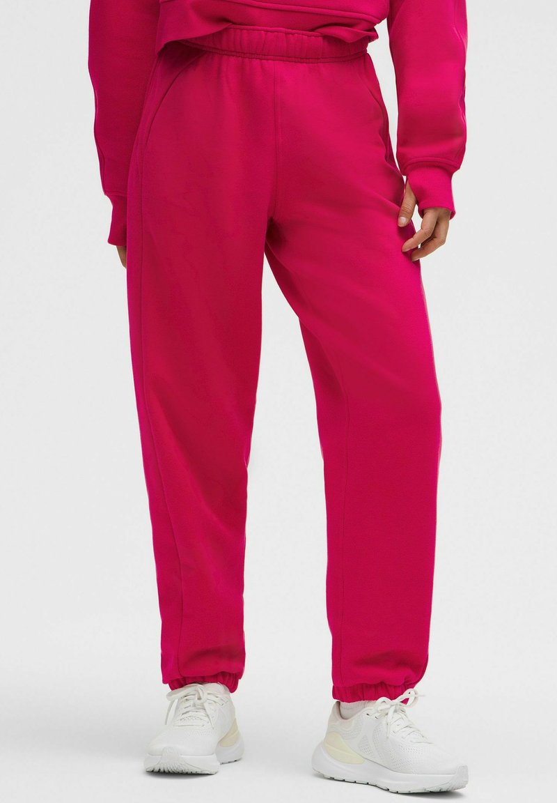 Fuchsia joggerbyxor med elastisk midja och muddar, tillverkade av ett slätt tyg. Parat med vita sportskor.