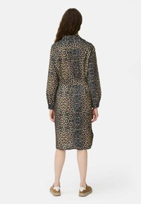 Frau mit langen Haaren, die ein tailliertes, langärmliges Kleid im Leopardenmuster und braune Sneakers trägt, steht mit dem Rücken zu einer weißen Hintergrund.