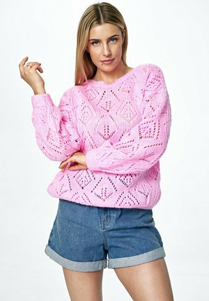 Różowy sweter dzianinowy z otwartym wzorem diamentowym, długimi rękawami i ściągaczami, w połączeniu z dżinsowymi szortami.