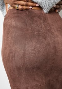 Des mains avec une bague en œil de tigre reposant sur la taille d'une personne portant une jupe en daim marron et un pull gris clair.